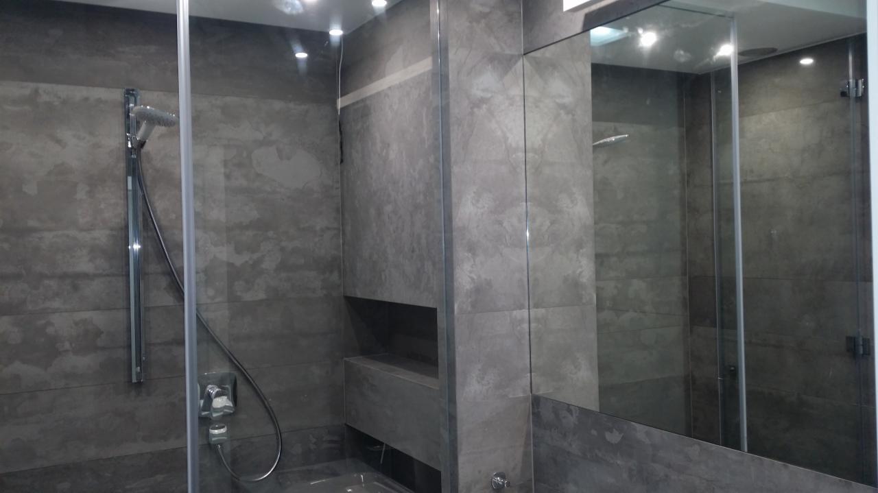 Szara łazienka z kabiną prysznicową typu walk-in, płytki imitujące beton, wbudowana półka w ścianie.