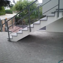Zewnętrzne schody betonowe z szarą, metalową balustradą prowadzące do budynku, ułożone na szarej kostce brukowej. Widoczny fragment ogrodu i ogrodzenia w tle.