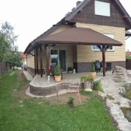 Dom z jasnej cegły z zadaszonym tarasem, widoczne elementy ogrodu: skalniak z małym strumykiem, trawnik z koniczyną, w tle ogrodzenie z brązowych desek.