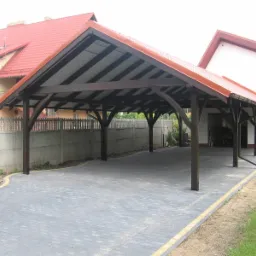 Zadaszona wiata garażowa z ciemnego drewna, kryta blachodachówką w kolorze czerwonym, na posesji z kostką brukową i betonowym ogrodzeniem.