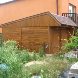 Drewniany budynek gospodarczy z dwuspadowym dachem z brązowej dachówki bitumicznej, przylegający do pomarańczowego domu z balkonem, otoczony bujną roślinnością, w tym winoroślą i wysoką trawą.