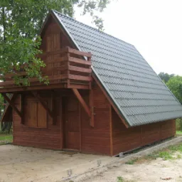 Mały drewniany domek z balkonem na tle zieleni, pokryty szarą blachodachówką, z widoczną konstrukcją wsporczą balkonu i betonową obwódką wokół budynku.