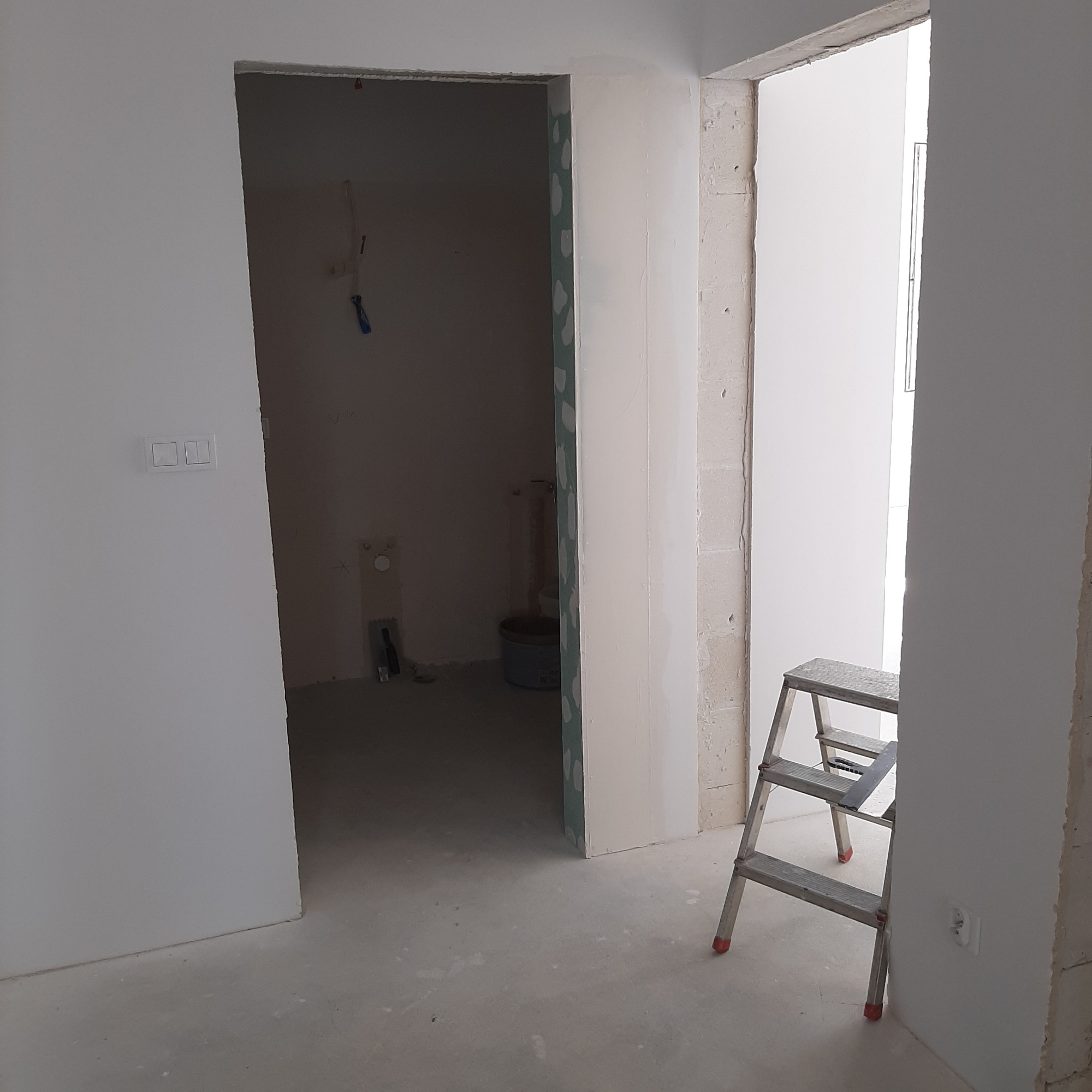 Wnętrze w trakcie prac wykończeniowych: widoczna drabina malarska, ściany po nałożeniu gładzi, przygotowane do malowania, w tle pomieszczenie łazienki z widocznymi przyłączami wodno-kanalizacyjnymi.