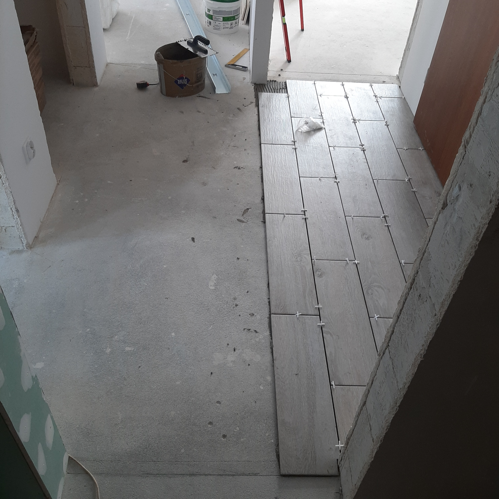 Układanie płytek podłogowych imitujących drewno w trakcie prac wykończeniowych, widoczny kubeł z klejem, poziomica oraz krzyżyki dystansowe.