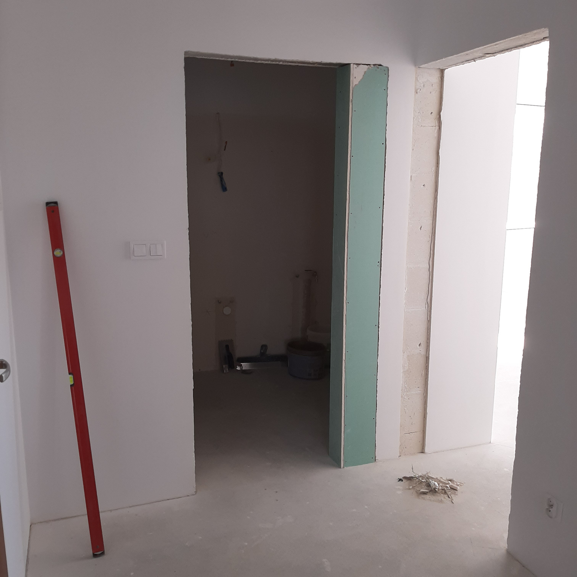 Wnętrze w trakcie remontu: widok na surową łazienkę przez otwór drzwiowy z zamontowaną płytą gipsowo-kartonową, czerwona poziomica oparta o ścianę, widoczne instalacje i narzędzia w tle.
