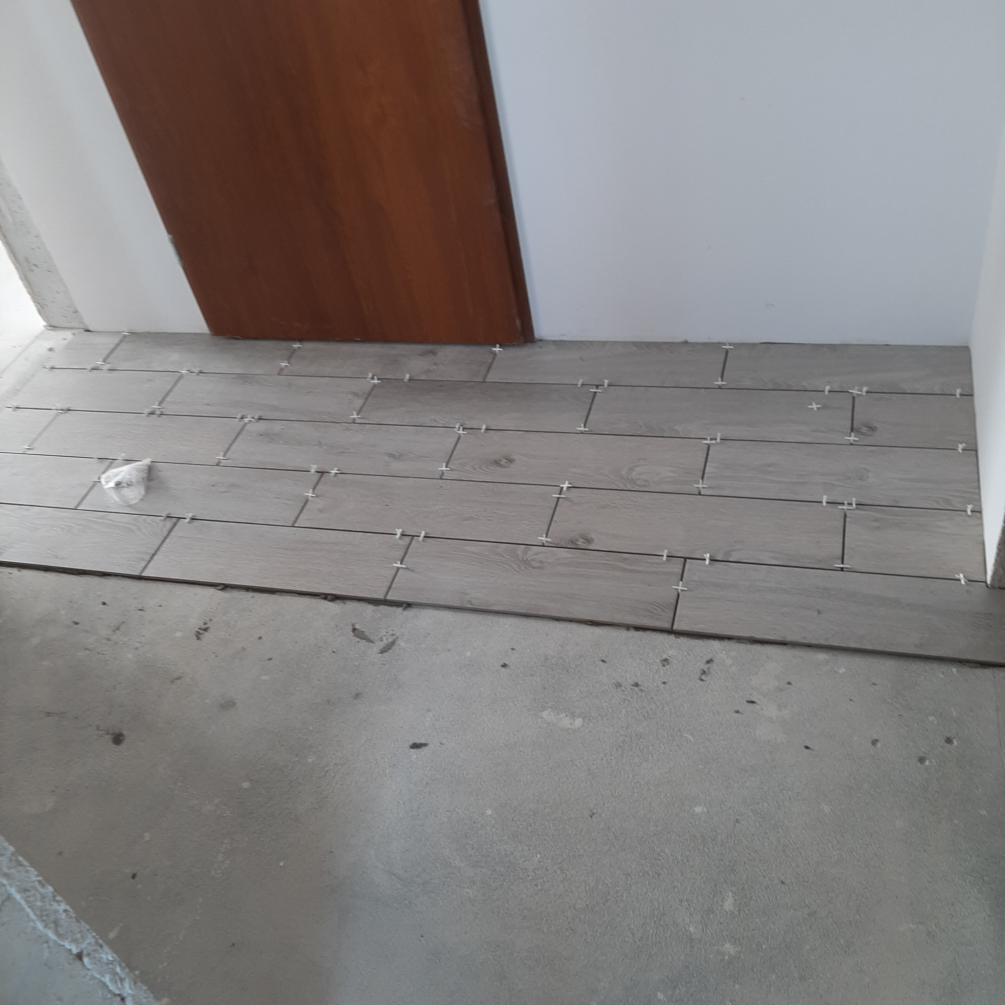 Układanie podłogi z płytek imitujących drewno w łazience, widoczne krzyżyki dystansowe i niezagruntowana podłoga, brązowe drzwi w tle.