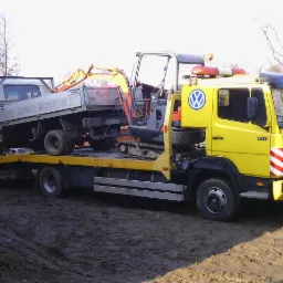 Żółty samochód ciężarowy z lawetą transportuje szarą ciężarówkę i pomarańczową mini-koparkę na tle drzew bez liści i ziemi.
