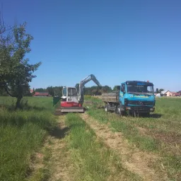Mini koparka Takeuchi TB53FR załadowuje ziemię do wywrotki Mercedes-Benz na tle zielonego pola i błękitnego nieba.