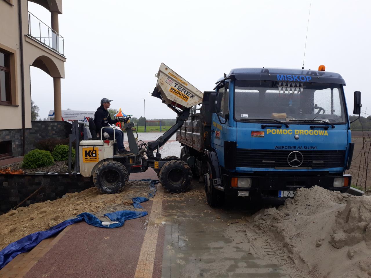 Operator minikoparki Terex załadowuje ziemię na ciężarówkę MISKOP z napisem 'Pomoc Drogowa' na tle budynku.