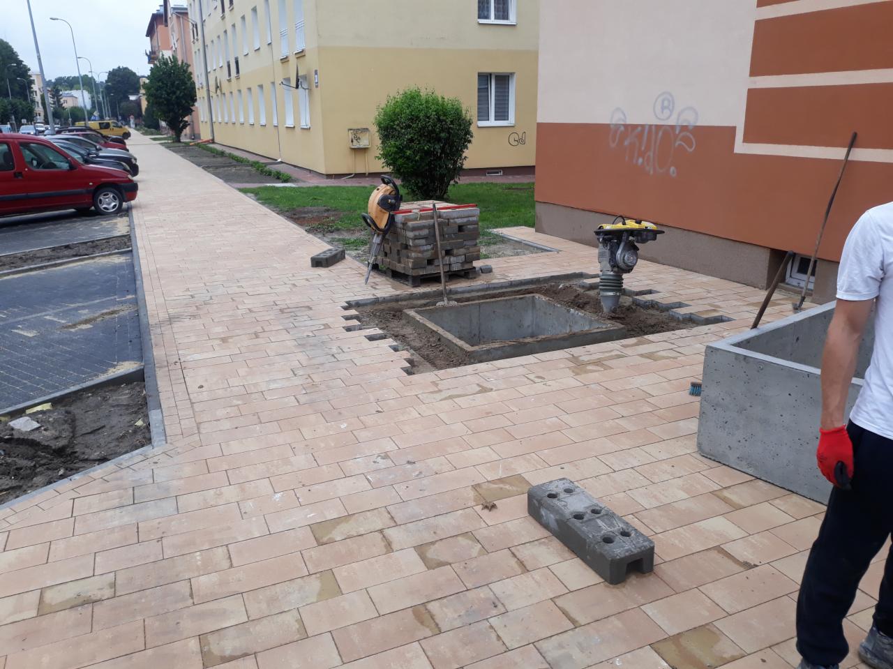 Układanie kostki brukowej na chodniku, widoczna ubijarka wibracyjna, paleta z kostką oraz betonowy element, prawdopodobnie studzienka kanalizacyjna.