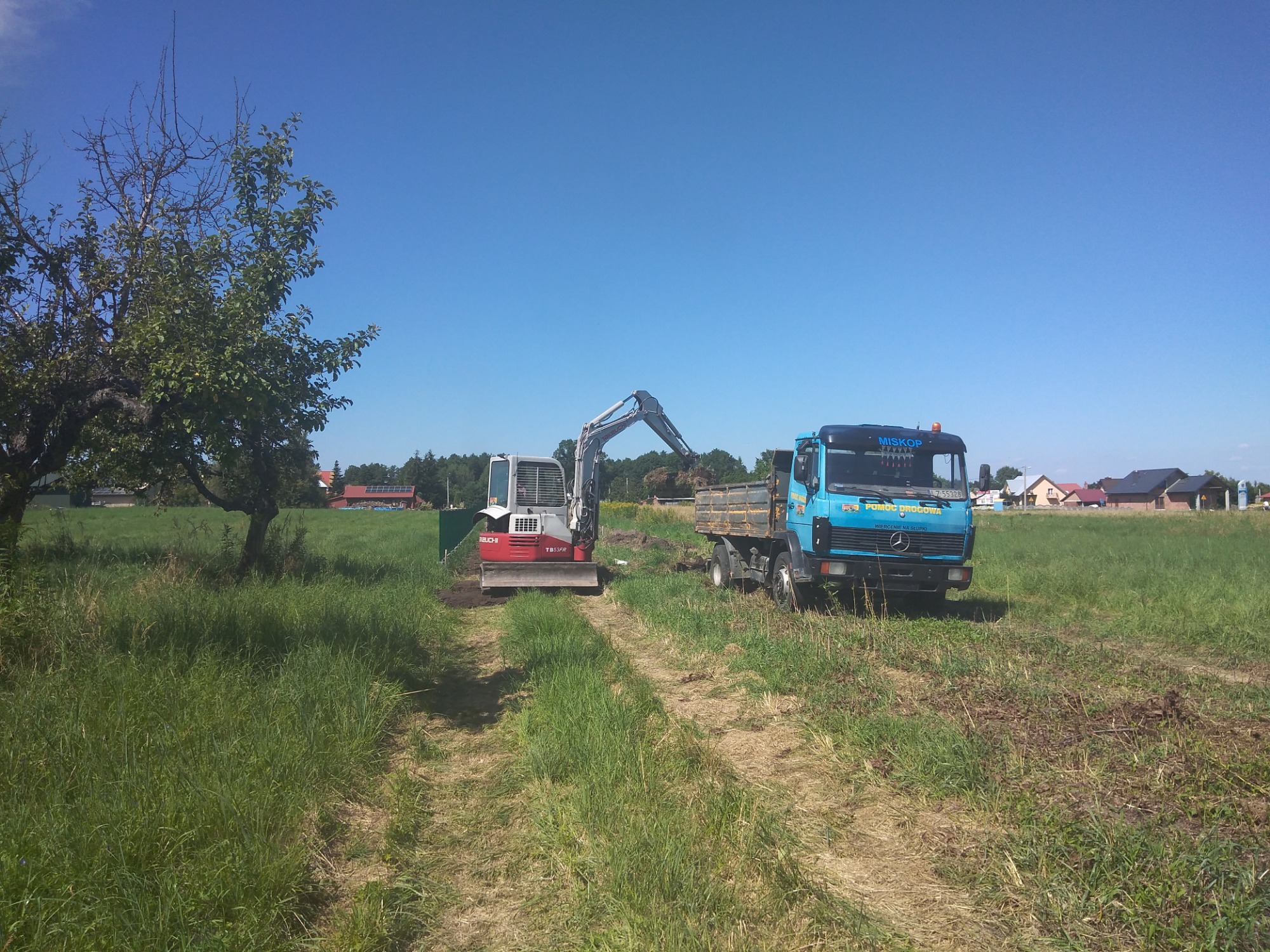 Mini koparka Takeuchi TB53FR załadowuje ziemię do wywrotki Mercedes-Benz na tle zielonego pola i błękitnego nieba.