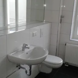 Wykończona łazienka w kamienicy z białą umywalką, toaletą i czarną podłogą z płytek, widoczne białe płytki na ścianach i duże lustro.