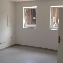 Pusty pokój po adaptacji, z szarymi płytkami podłogowymi, białymi ścianami, dwoma oknami z widokiem na konstrukcję budynku i wystającym przewodem z sufitu.