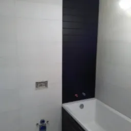 Wykończona łazienka z białą wanną i pionowym pasem czarnych płytek na ścianie, widoczne przyłącza wodne i elektryczne.