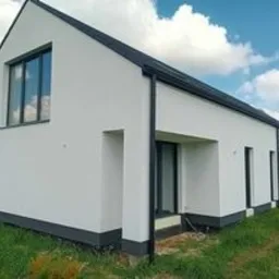 Nowoczesny, parterowy dom z białymi ścianami elewacji, czarnymi ramami okiennymi i dachem, widoczny w słoneczny dzień na tle błękitnego nieba z chmurami.