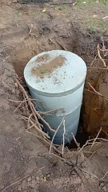 Betonowy zbiornik cylindryczny w wykopie ziemnym, widoczne wystające korzenie drzew, ziemia na pokrywie zbiornika.