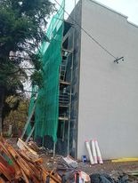 Budynek w trakcie prac elewacyjnych, osłonięty zieloną siatką rusztowania, widoczne materiały izolacyjne i pozostałości po pracach budowlanych.