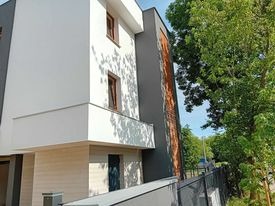 Nowoczesny, trzypiętrowy budynek z elewacją w kolorach białym, szarym i pomarańczowym, z widocznym balkonem i oknami z brązowymi ramami. Częściowo zacieniony przez drzewo.