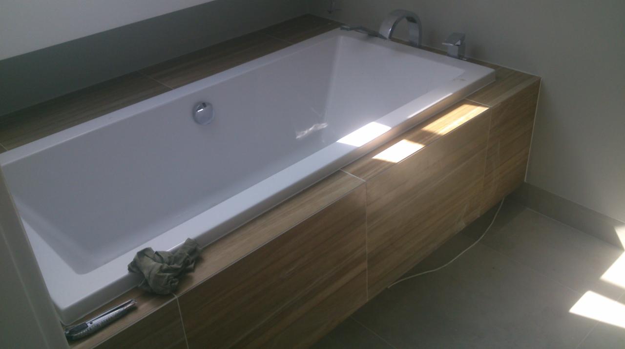 Biała wanna z obudową z płytek imitujących drewno, nowoczesna armatura, szara podłoga i ściany. Fragment nożyka i szmatki na obudowie wanny.