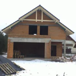 Dom w stanie surowym otwartym, z widocznym zbrojonym nadprożem nad garażem i stertą materiałów budowlanych na ośnieżonym terenie. Budowa w zimowej scenerii.