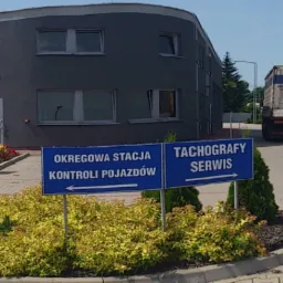 Budynek Okręgowej Stacji Kontroli Pojazdów z tablicami informacyjnymi i zaparkowaną ciężarówką w tle, otoczony zielenią i kwiatami.