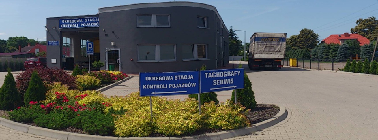 Budynek Okręgowej Stacji Kontroli Pojazdów z tablicami informacyjnymi i zaparkowaną ciężarówką w tle, otoczony zielenią i kwiatami.