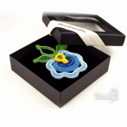 Ręcznie robiona biżuteria sutasz w kształcie niebieskiego kwiatu z zielonymi listkami, zapakowana w czarne pudełko z okienkiem, wykonana przez firmę z Gdańska.