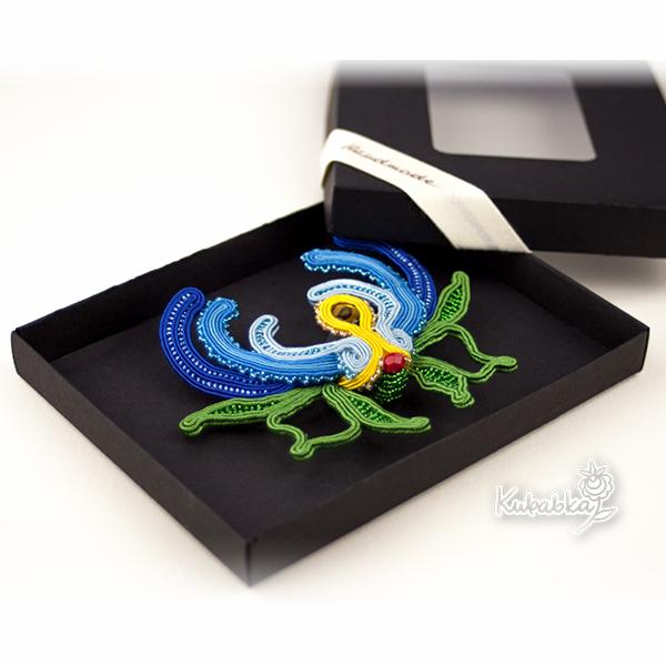 Ręcznie robiona biżuteria sutasz w kształcie kwiatu, z niebieskimi, żółtymi i zielonymi sznurkami, ozdobiona czerwonym koralikiem, prezentowana w czarnym pudełku z okienkiem.