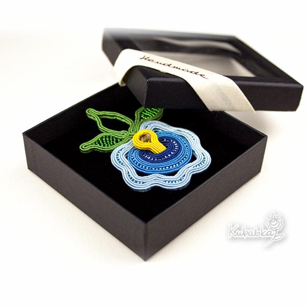 Ręcznie robiona biżuteria sutasz w kształcie niebieskiego kwiatu z zielonymi listkami, zapakowana w czarne pudełko z okienkiem, wykonana przez firmę z Gdańska.