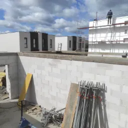 Budowa domu z białych bloczków: ściany, rusztowania, podpory. W tle nowoczesne budynki i pracownik na rusztowaniu. Widok z góry na plac budowy w słoneczny dzień.