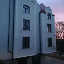 Dwupiętrowy dom murowany z dachem pokrytym czerwoną dachówką, widziany z dołu pod kątem, z widoczną elewacją w kolorze jasnoszarym i ozdobnymi detalami architektonicznymi, na tle wieczornego nieba...