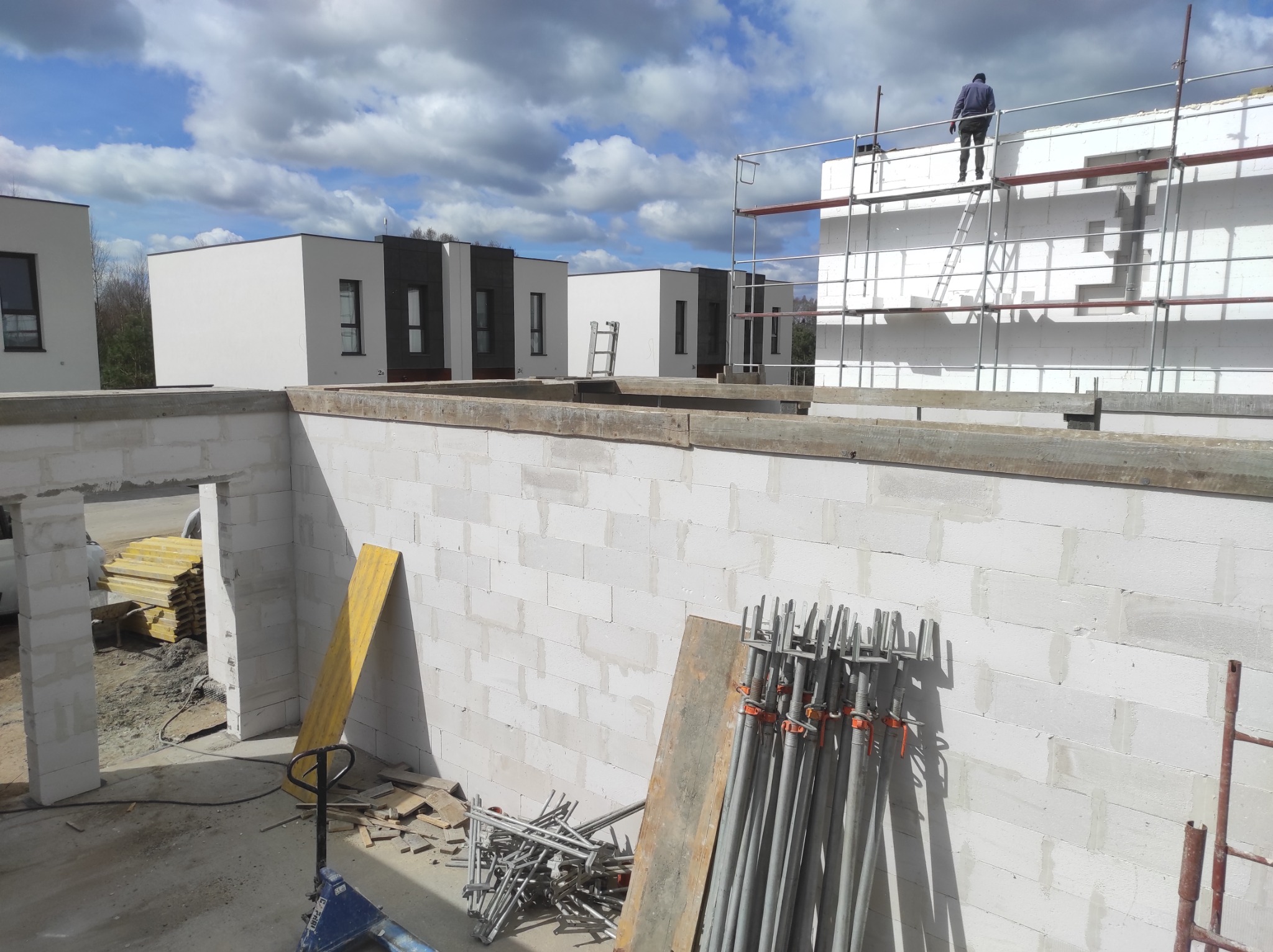 Budowa domu z białych bloczków: ściany, rusztowania, podpory. W tle nowoczesne budynki i pracownik na rusztowaniu. Widok z góry na plac budowy w słoneczny dzień.