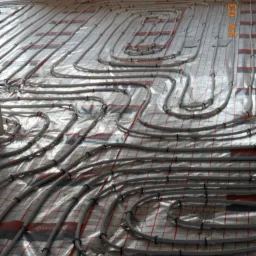 Układ rur ogrzewania podłogowego na folii izolacyjnej z rastrem. Widoczne mocowania rur. Ujęcie z góry, z datą realizacji w prawym górnym rogu.