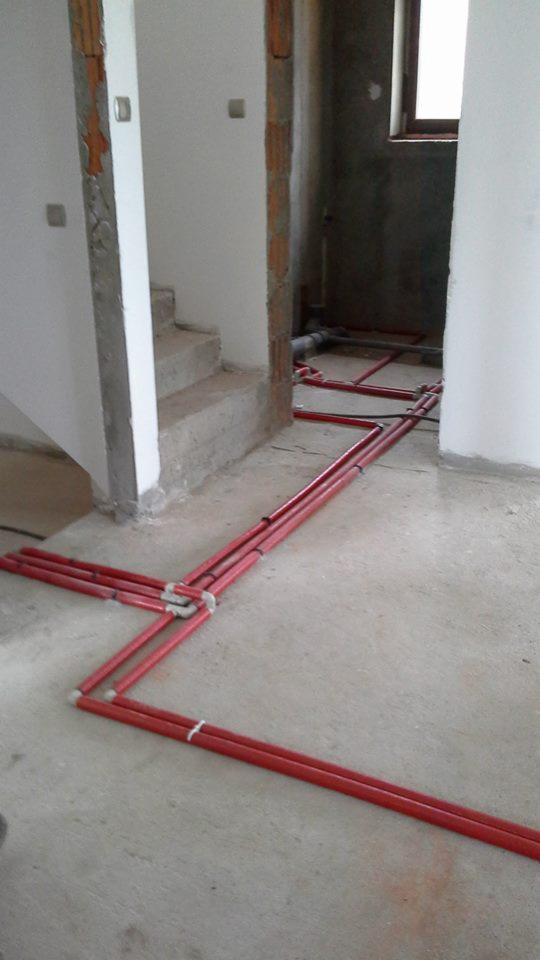 Czerwone rury instalacyjne ułożone na surowym betonowym podłożu w trakcie montażu w nowym budynku, widoczne fragmenty schodów i ściany w stanie deweloperskim.