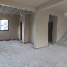 Tynki Bielsko Łukasz Kiecza - Surowe wnętrze domu w trakcie prac wykończeniowych, ściany pokryte świeżym tynkiem, widoczne instalacje elektryczne i hydrauliczne, surowy betonowy strop, schody w stanie surowym.