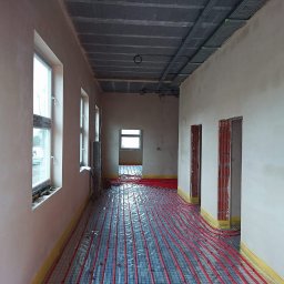 Tynki Bielsko Łukasz Kiecza - Wnętrze budynku w trakcie prac wykończeniowych: ściany po nałożeniu jasnego tynku, instalacja ogrzewania podłogowego rozłożona na podłodze, widoczne otwory drzwiowe z elementami cegieł i instalacje...