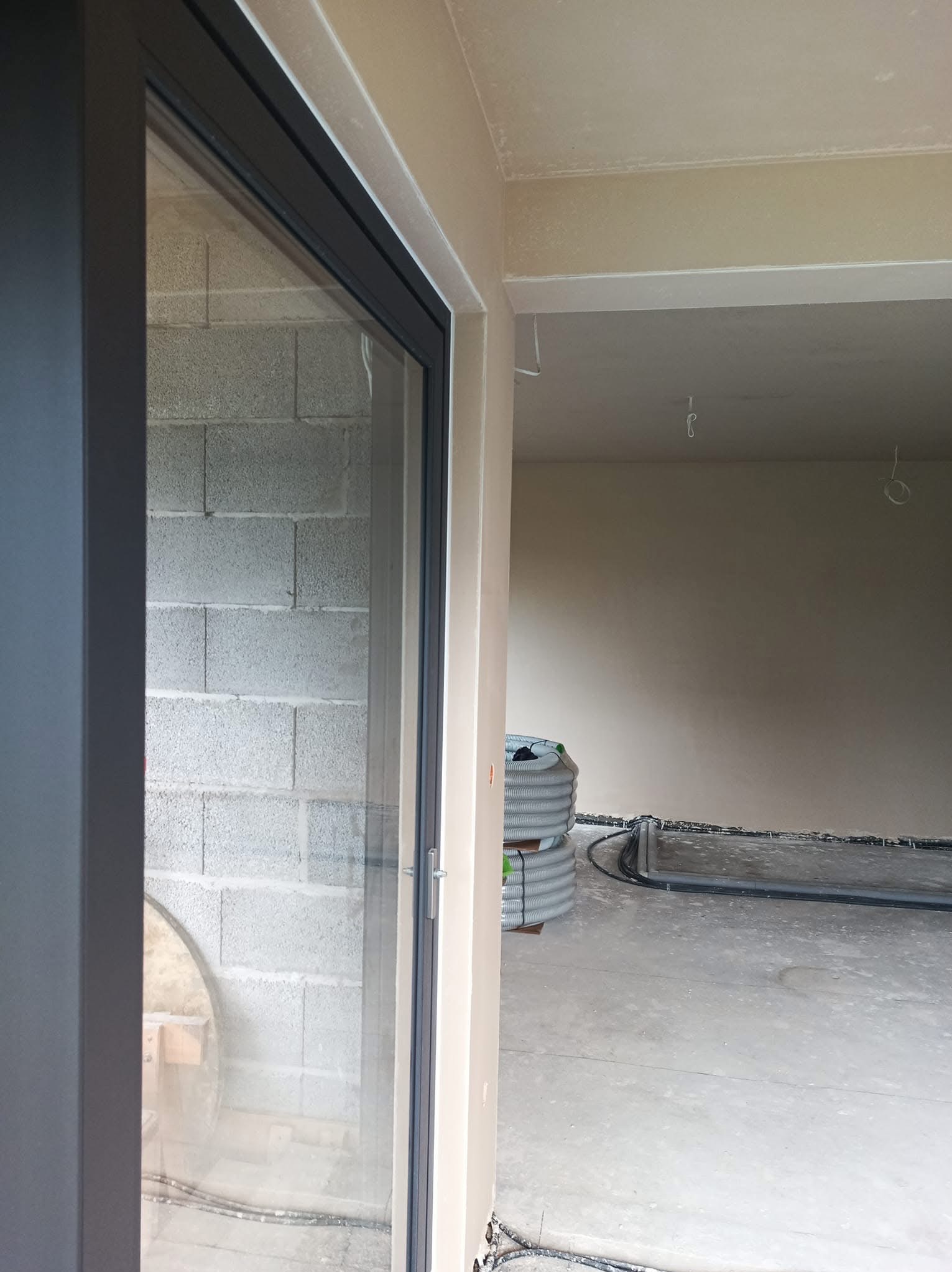Wnętrze w stanie deweloperskim z widocznymi szarymi ścianami z bloczków, częściowo otynkowanymi ścianami w odcieniach beżu, zwiniętymi rurami wentylacyjnymi i instalacjami elektrycznymi na podłodze.