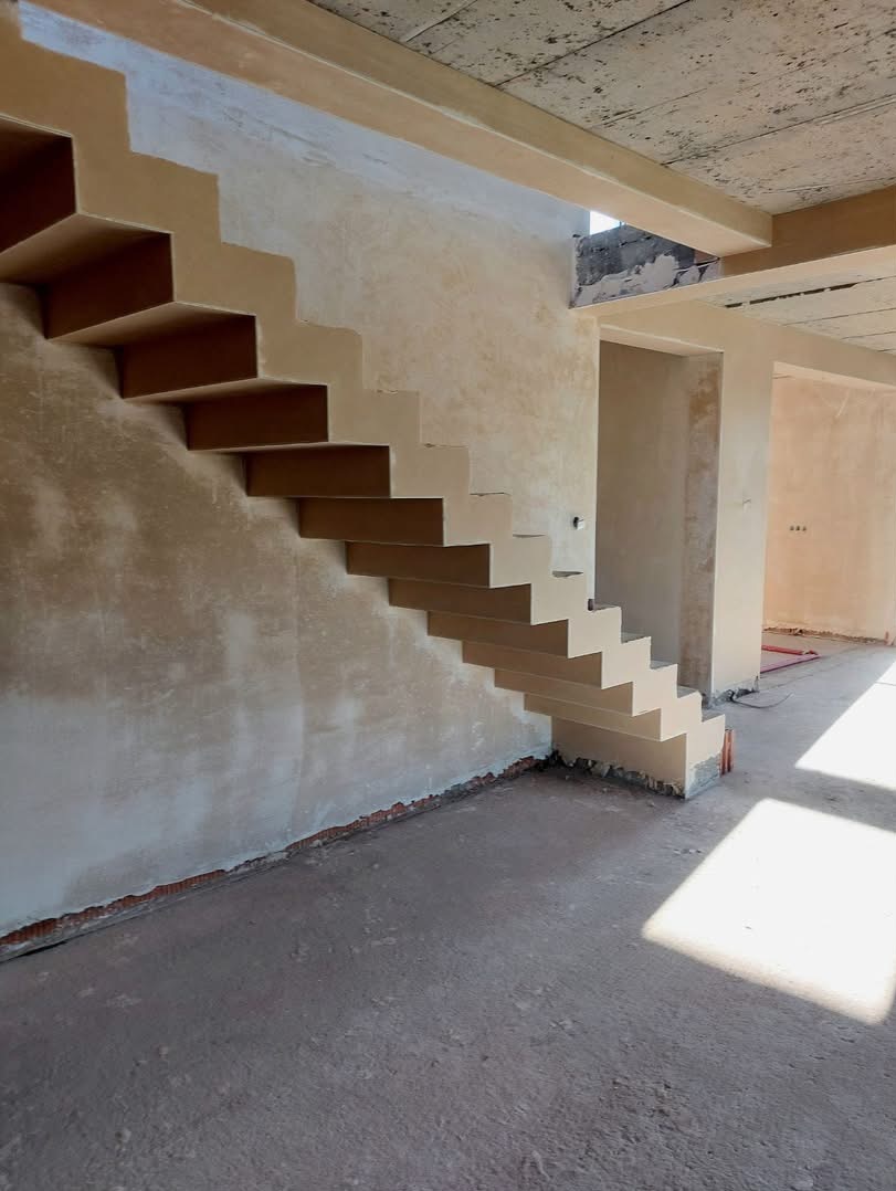Surowe schody wspornikowe z nałożonymi jasnymi tynkami gipsowymi na ścianach w stanie deweloperskim, widoczne ślady po cegłach przy podłodze, surowy betonowy sufit.