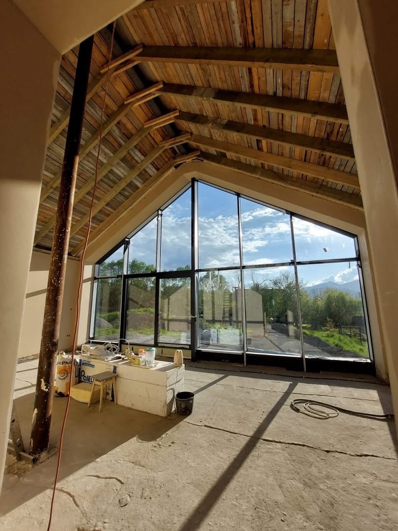 Wnętrze domu w trakcie tynkowania maszynowego, widoczne surowe ściany, drewniana konstrukcja dachu, duże okno z widokiem na zieleń i góry w tle, oraz narzędzia i materiały budowlane.