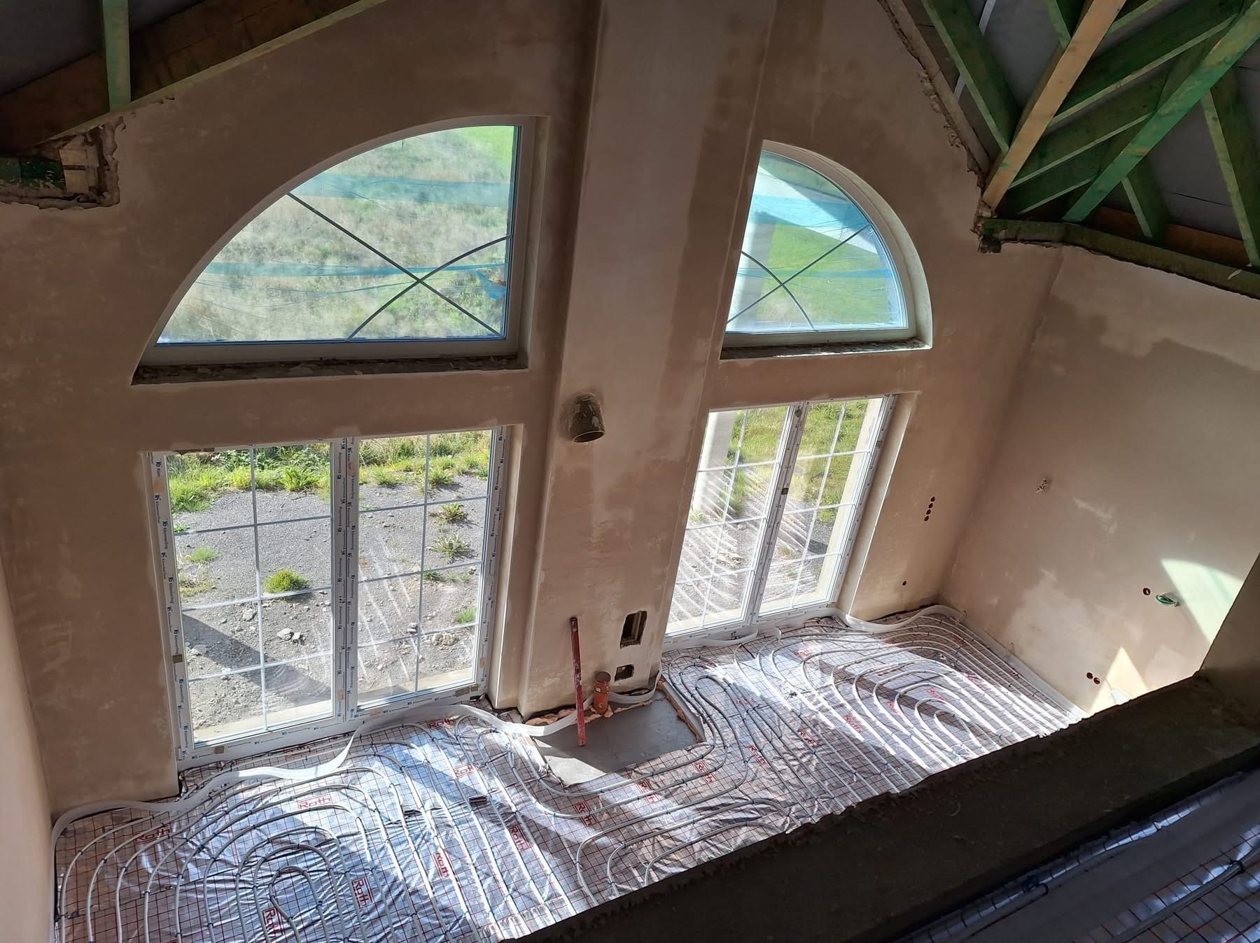 Wnętrze w stanie surowym z widoczną instalacją ogrzewania podłogowego, świeżo położonymi tynkami i nietypowymi oknami w szczycie budynku.