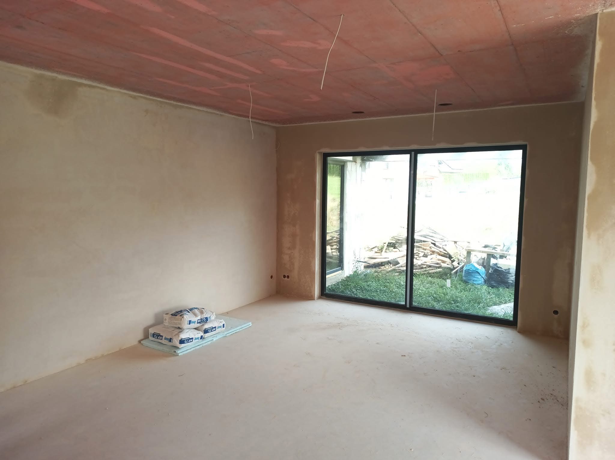 Świeżo otynkowane ściany w pomieszczeniu w Bielsku-Białej, widoczne worki z zaprawą i duże okno przesuwne z widokiem na ogród.