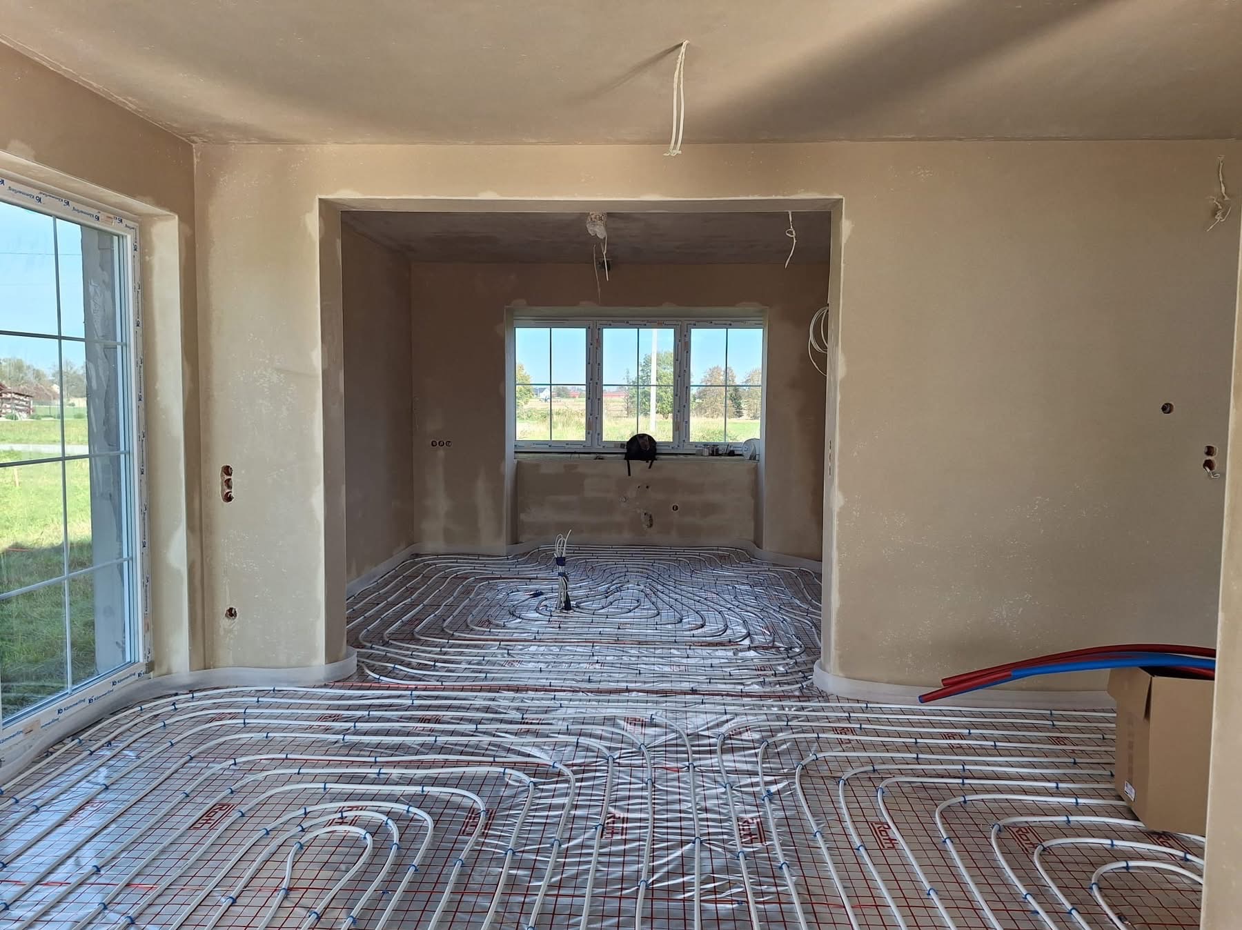 Wnętrze domu w trakcie prac wykończeniowych: ściany po nałożeniu gładzi gipsowej, widoczne instalacje ogrzewania podłogowego na siatce, okno z widokiem na zieleń.