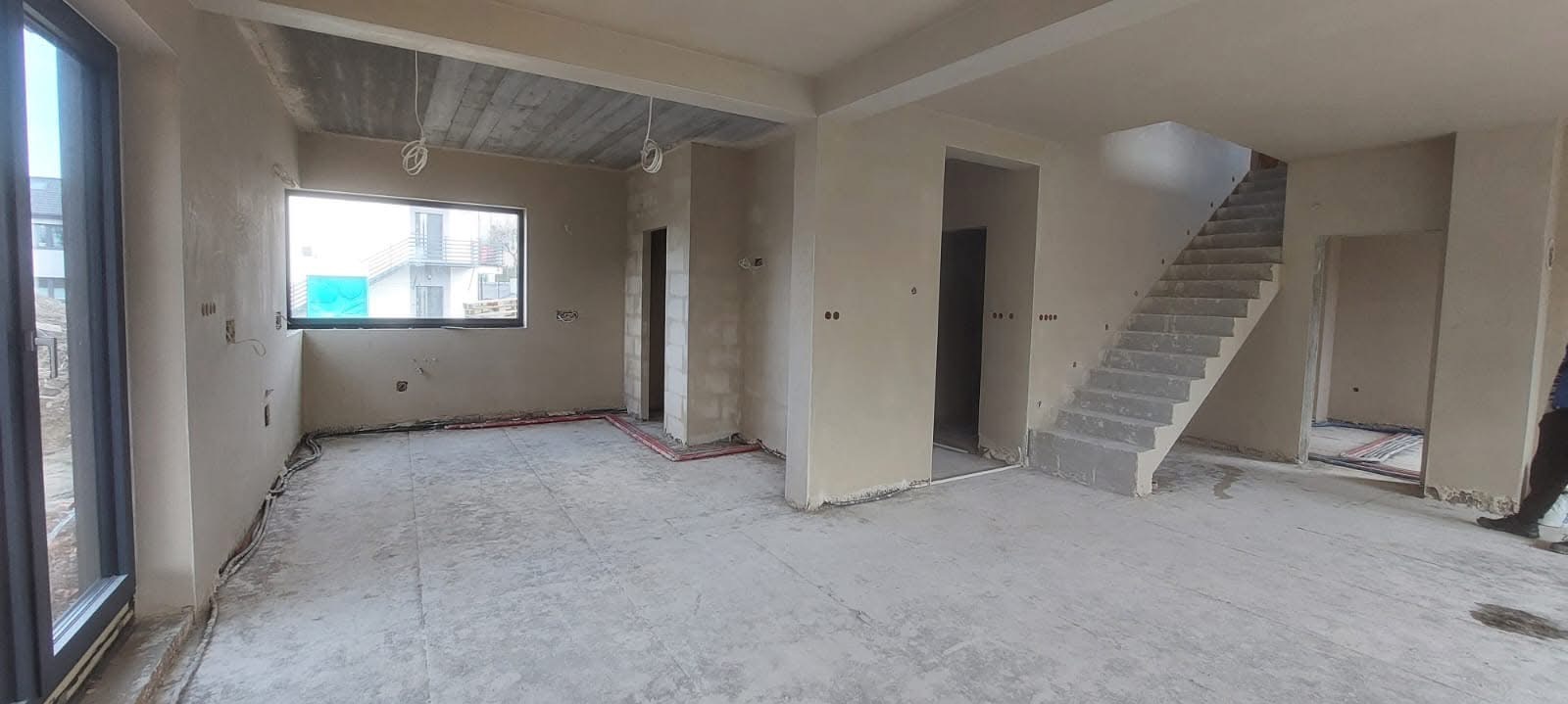 Surowe wnętrze domu w trakcie prac wykończeniowych, ściany pokryte świeżym tynkiem, widoczne instalacje elektryczne i hydrauliczne, surowy betonowy strop, schody w stanie surowym.