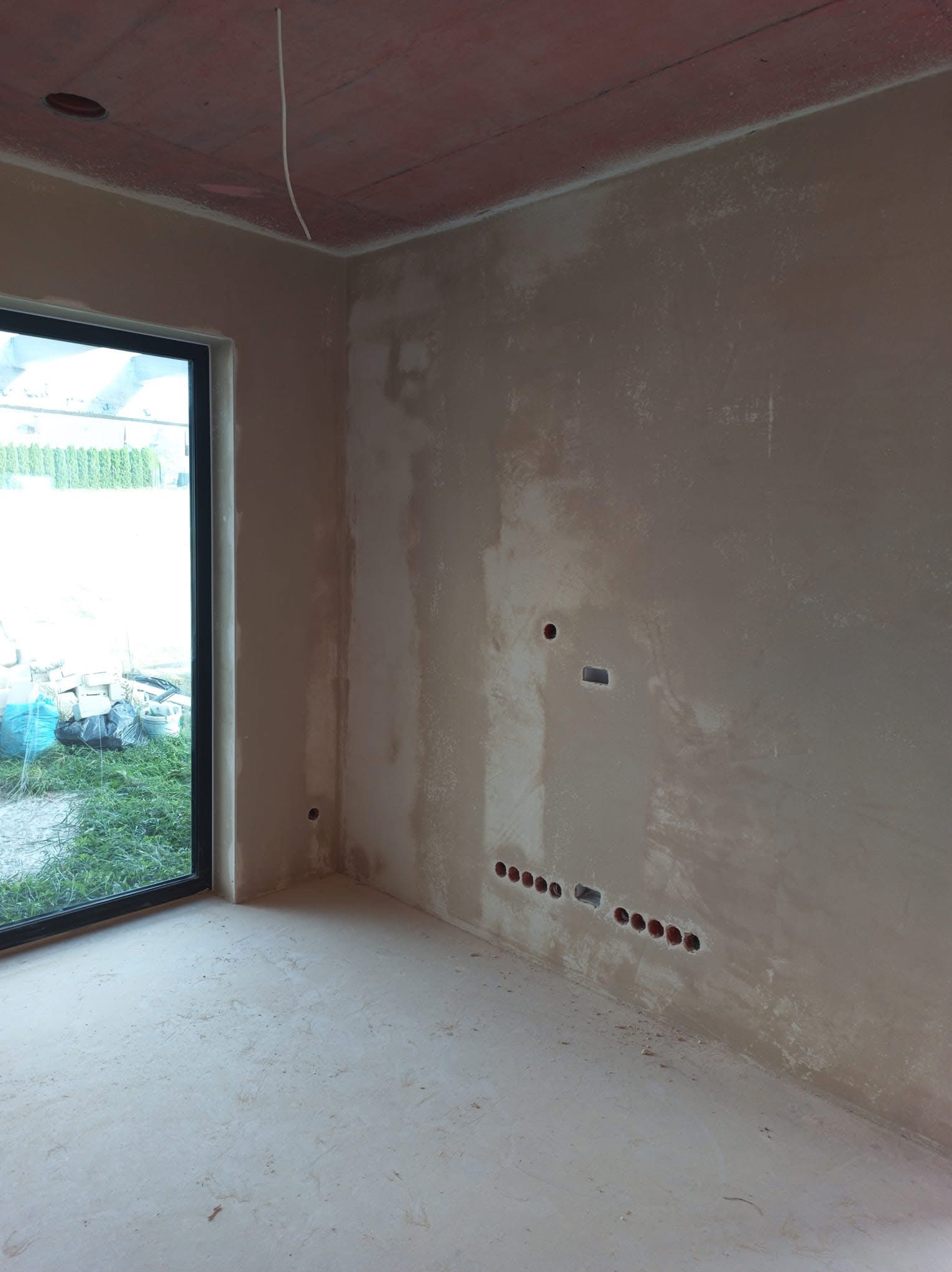 Świeżo nałożone tynki maszynowe na ścianach wewnętrznych budynku w stanie surowym, widoczne otwory pod gniazdka elektryczne i wystający kabel z sufitu, przez okno widoczny ogród.