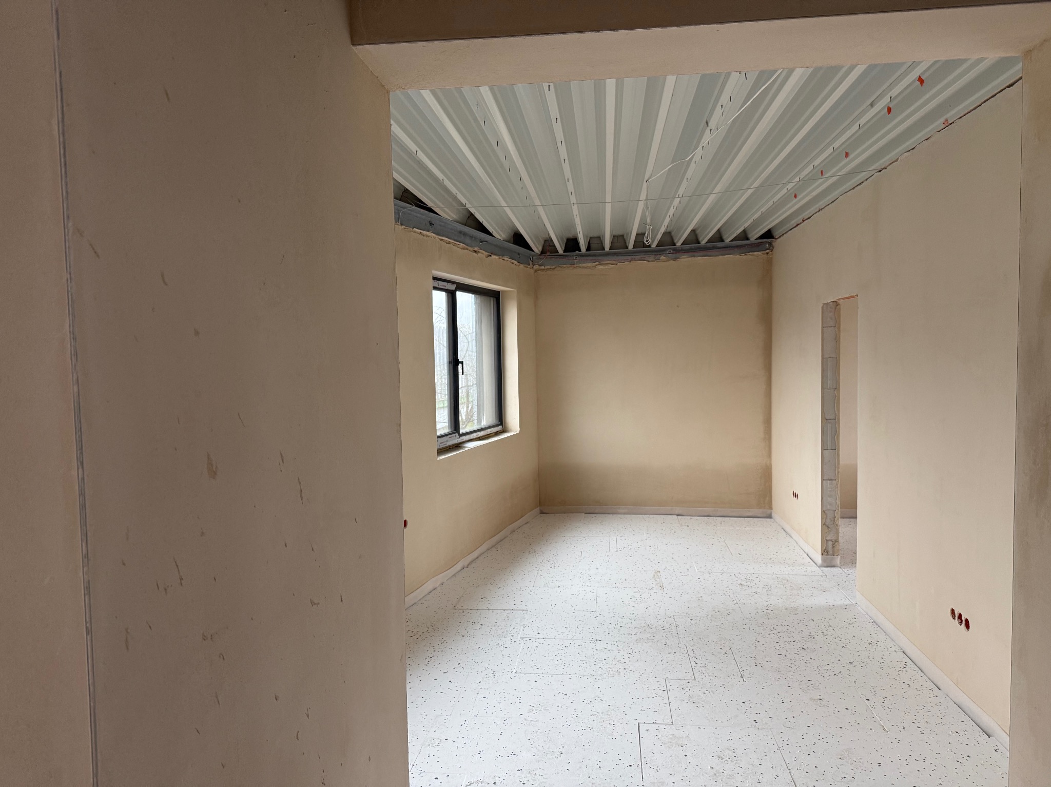 Wnętrze w stanie deweloperskim po nałożeniu jasnych tynków, widoczne gładkie ściany, sufit z widoczną konstrukcją i biała posadzka z drobnym wzorem.