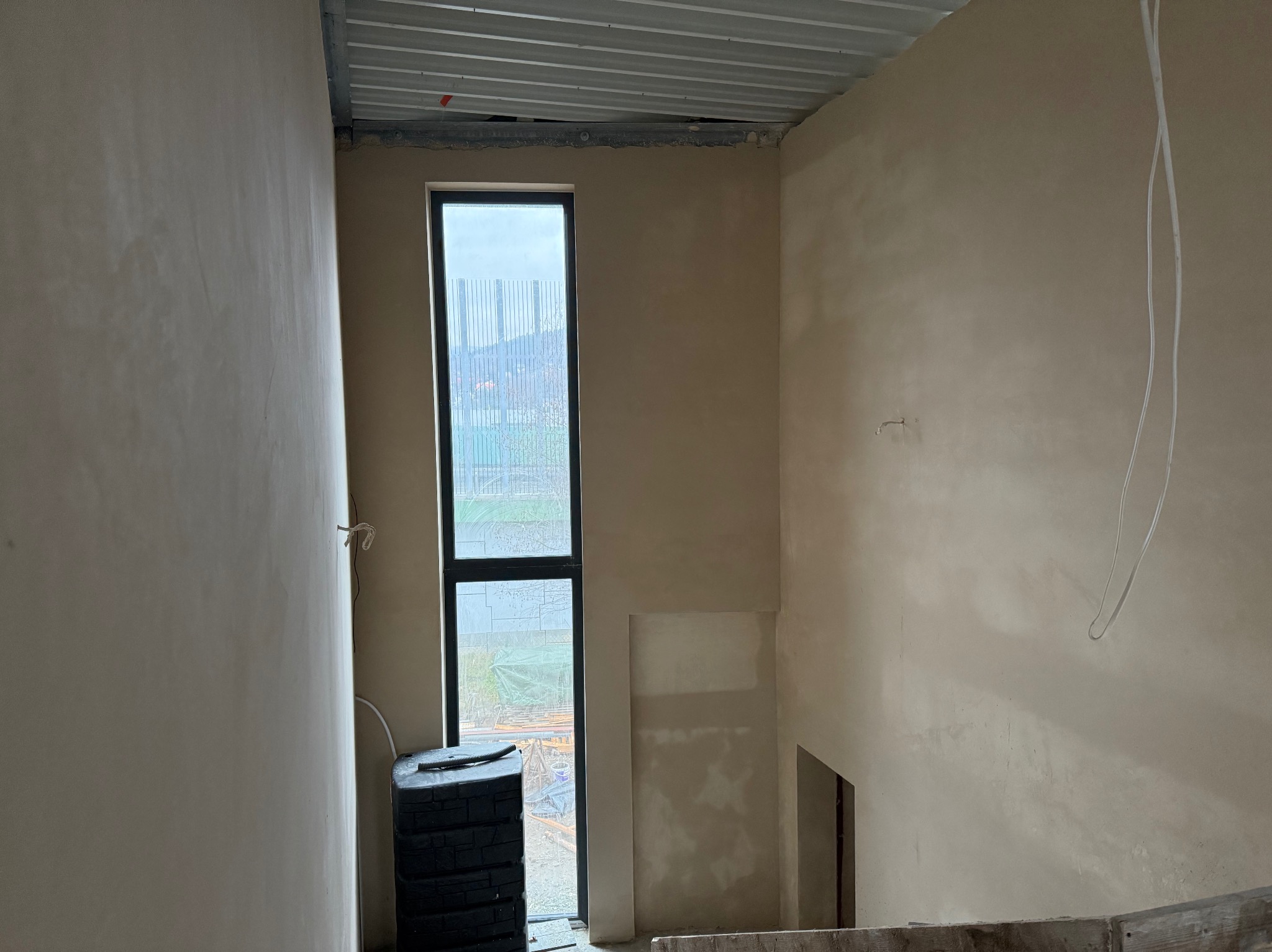 Wnętrze w trakcie tynkowania maszynowego z widocznym oknem, stertą czarnych zbiorników na wodę i surowym, metalowym stropem.