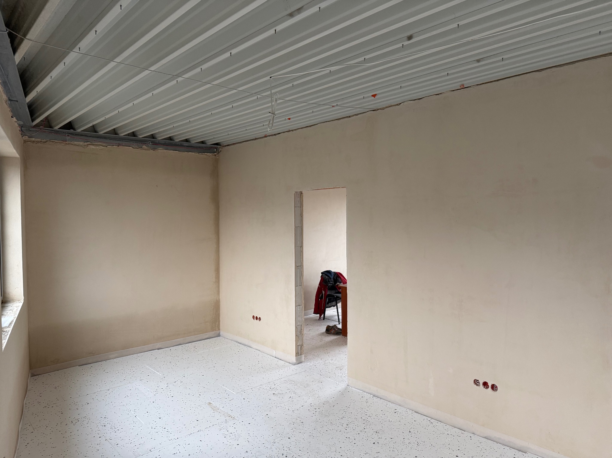 Wnętrze pokoju po nałożeniu gładzi, widoczne gniazdka elektryczne, podłoga zabezpieczona folią, surowy sufit z blachy trapezowej i metalowymi profilami, w przejściu widoczne krzesło i ubrania.
