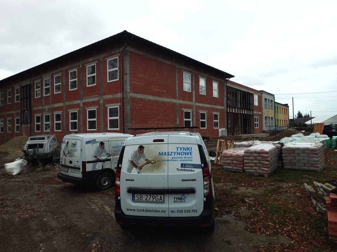 Budynek w stanie surowym otwartym z widocznymi dwoma samochodami dostawczymi z reklamą firmy wykonującej usługi tynkarskie oraz workami z zaprawą tynkarską na paletach.
