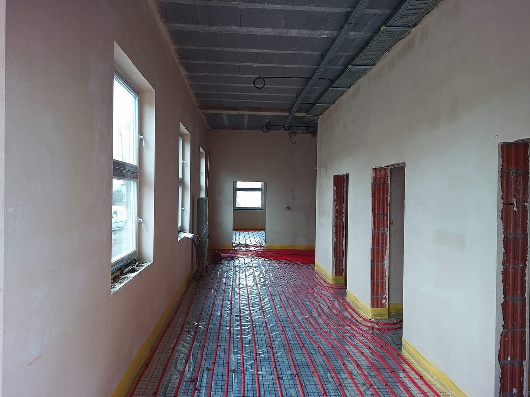 Wnętrze budynku w trakcie prac wykończeniowych: ściany po nałożeniu jasnego tynku, instalacja ogrzewania podłogowego rozłożona na podłodze, widoczne otwory drzwiowe z elementami cegieł i instalacje...