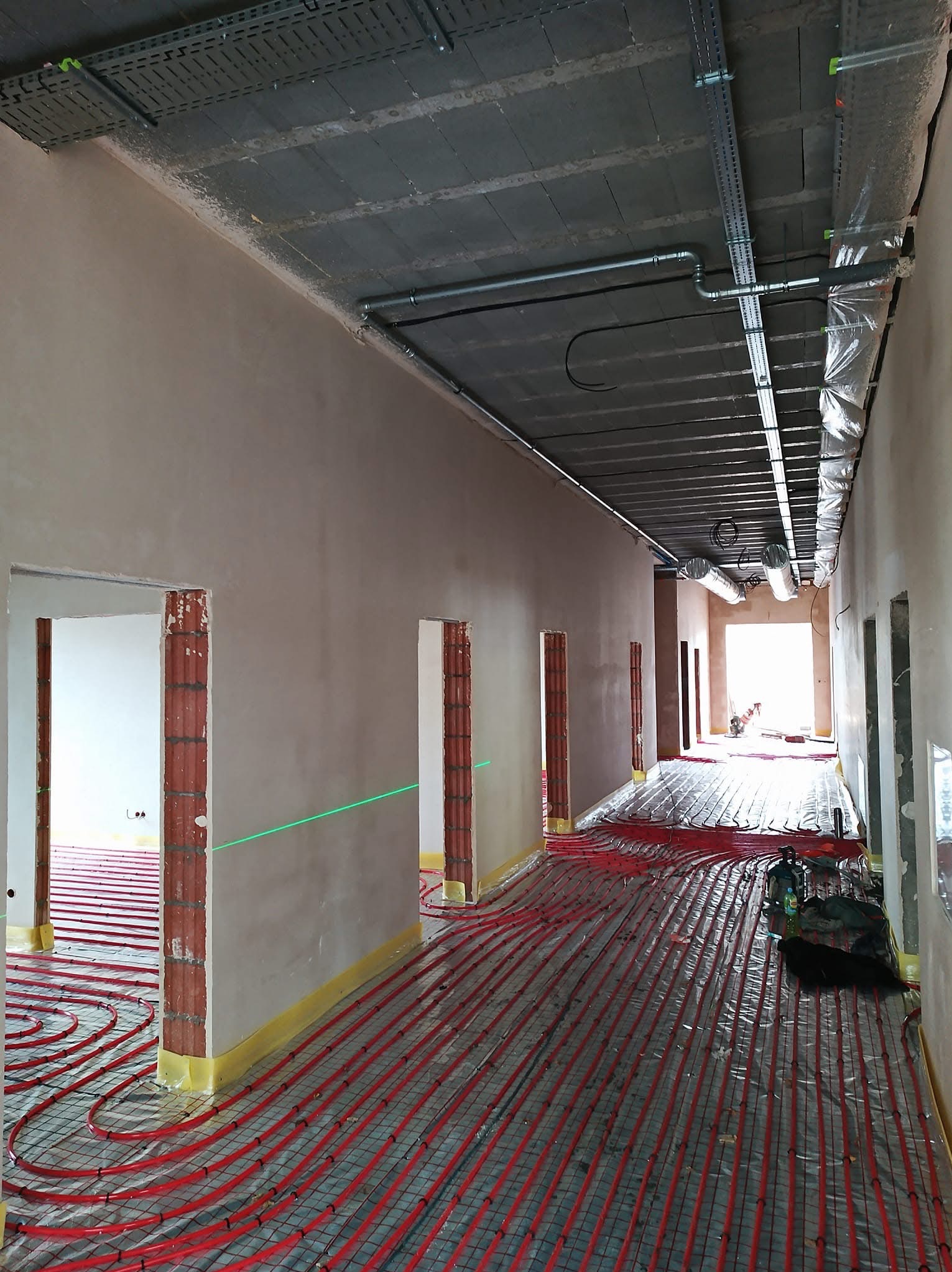 Korytarz w trakcie remontu: ściany po nałożeniu tynku, na podłodze rozłożone czerwone rury ogrzewania podłogowego, widoczny laserowy niwelator wyznaczający poziom.